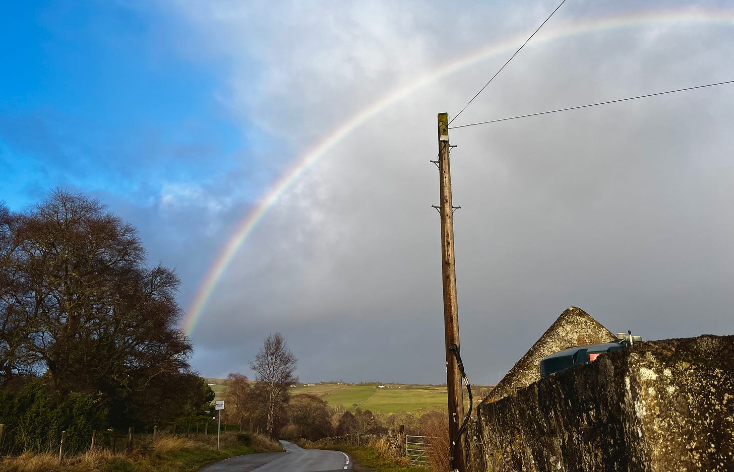 rainbow in a sky part-blue and part-deep gray, above a ruined stone building and distant hillside