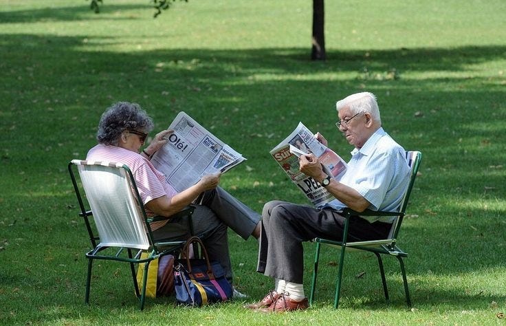 This may contain: two elderly people sitting in lawn chairs reading the paper on a sunny day with trees and grass behind them