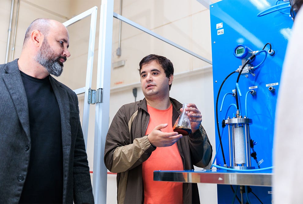 Duas pessoas em um laboratório ou espaço de trabalho técnico: à esquerda, pessoa com barba grisalha usando blazer cinza sobre camiseta preta; à direita, pessoa usando camiseta laranja sob jaqueta marrom, gesticulando com a mão direita enquanto segura um recipiente de vidro na mão esquerda, ambas posicionadas diante de um equipamento azul com tubagens, válvulas e display digital acoplados, com estrutura de vidro e metal ao fundo em ambiente com iluminação clara. Duas pessoas em um laboratório ou espaço de trabalho técnico: à esquerda, pessoa com barba grisalha usando blazer cinza sobre camiseta preta; à direita, pessoa usando camiseta laranja sob jaqueta marrom, gesticulando com a mão direita enquanto segura um recipiente de vidro na mão esquerda, ambas posicionadas diante de um equipamento azul com tubagens, válvulas e display digital acoplados, com estrutura de vidro e metal ao fundo em ambiente com iluminação clara.
