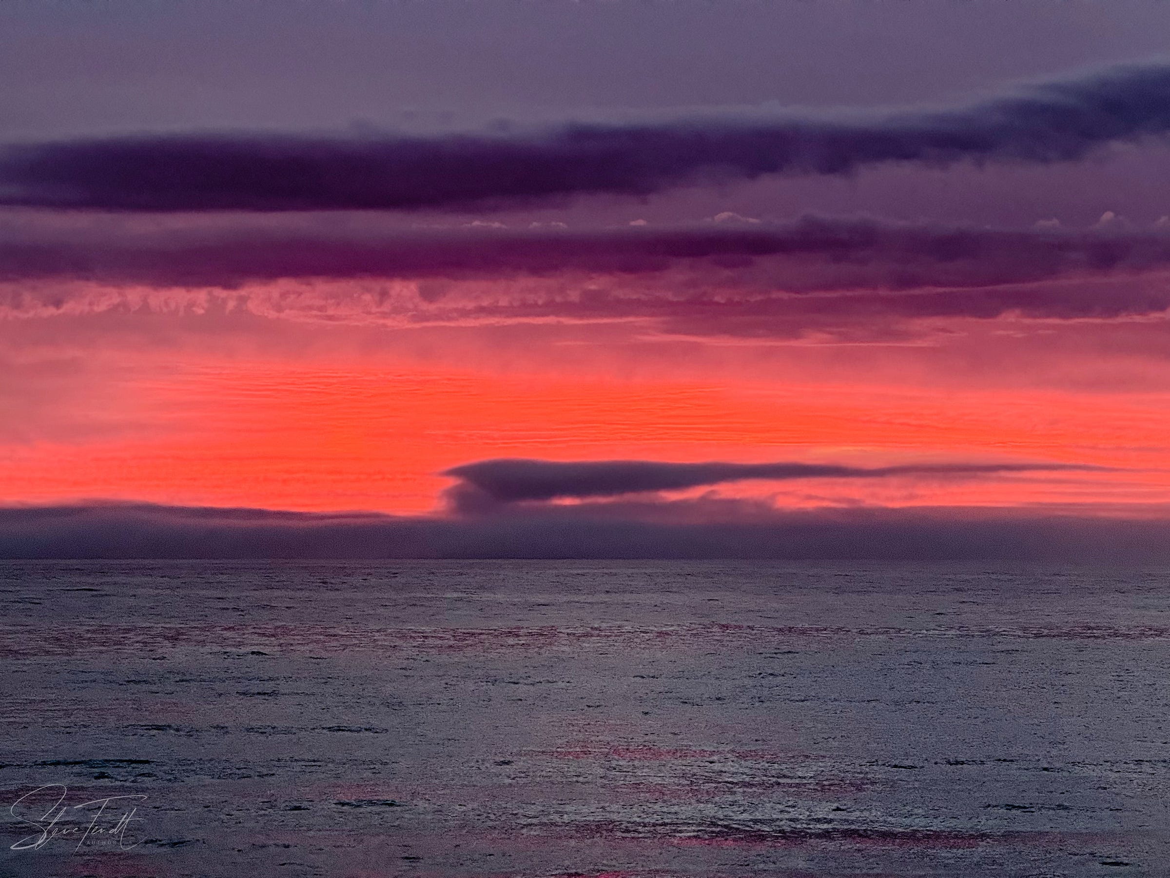 Photograph: spectacular red sunrise between dark grey-blue sea and clouds.