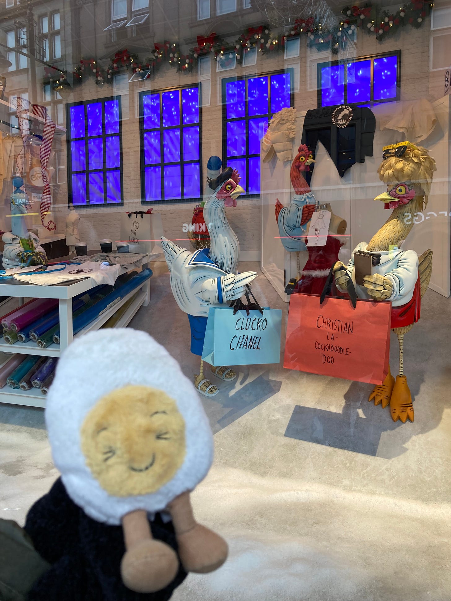 Photo of Dippy the soft toy egg outside of Fenwick's christmas window. It is a display of animatronic hens in a designer sewing room, with the hens wearing French-designer clothes and carrying bags that say Clucko Chanel and Christian La Cockadoodle-Doo. It is themed for the twelve days of Christmas, and this window is three French hens.
