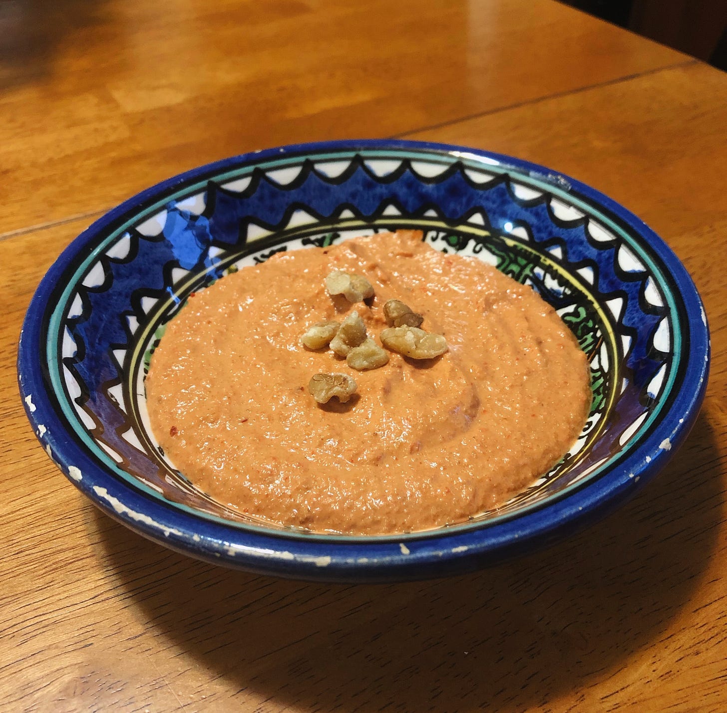 a light orange dip with hummus-like texture in a decorated ceramic bowl painted with blue designs. the dip is topped with chopped walnuts and the bowl is sitting on a table.