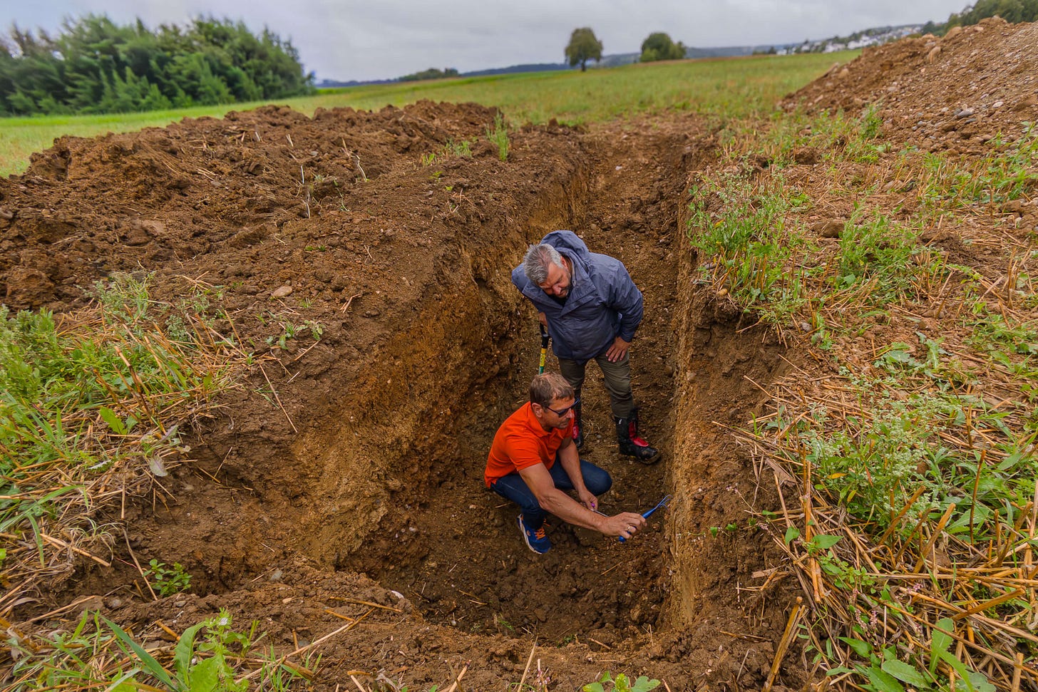 Die Landwirte Gerhard Weisshäupl und Sven Studer stehen in einer zwei Meter tiefen Bodenprofil-Grube auf einem Acker. Weisshäupl in orangem T-Shirt kniet und untersucht mit einem Werkzeug das Bodenprofil an der Grubenwand, Studer in blauer Jacke schaut interessiert zu. Rundherum liegen Erdhaufen, im Hintergrund sind Wiesen, Bäume und ein trüber Himmel zu sehen. Die Landwirte Gerhard Weisshäupl und Sven Studer stehen in einer zwei Meter tiefen Bodenprofil-Grube auf einem Acker. Weisshäupl in orangem T-Shirt kniet und untersucht mit einem Werkzeug das Bodenprofil an der Grubenwand, Studer in blauer Jacke schaut interessiert zu. Rundherum liegen Erdhaufen, im Hintergrund sind Wiesen, Bäume und ein trüber Himmel zu sehen.