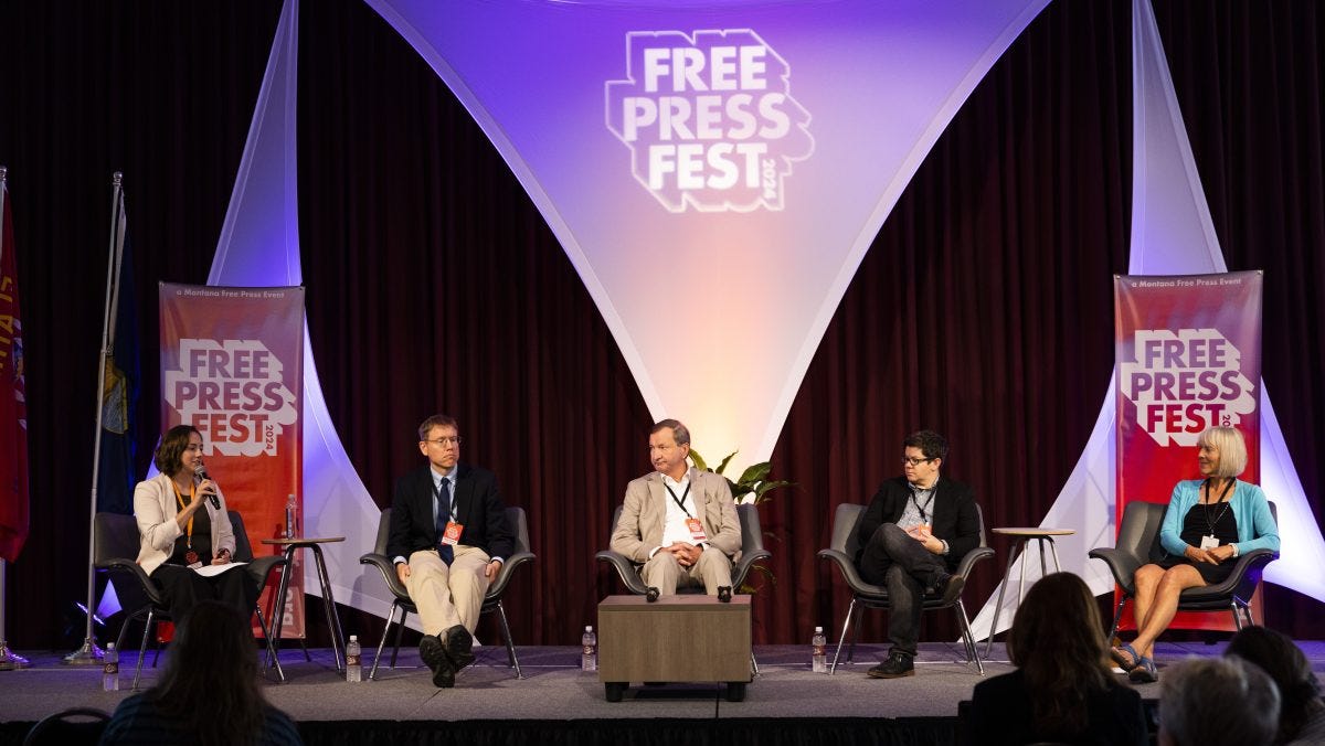 Five panelists on stage at Free Press Fest, seated under a large backdrop with festival logos and banners. Five panelists on stage at Free Press Fest, seated under a large backdrop with festival logos and banners.