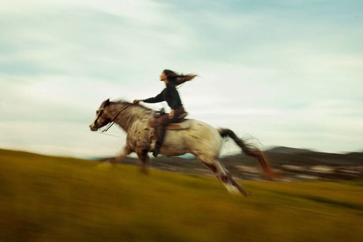 This may contain: a woman riding on the back of a white horse through a green grass covered field