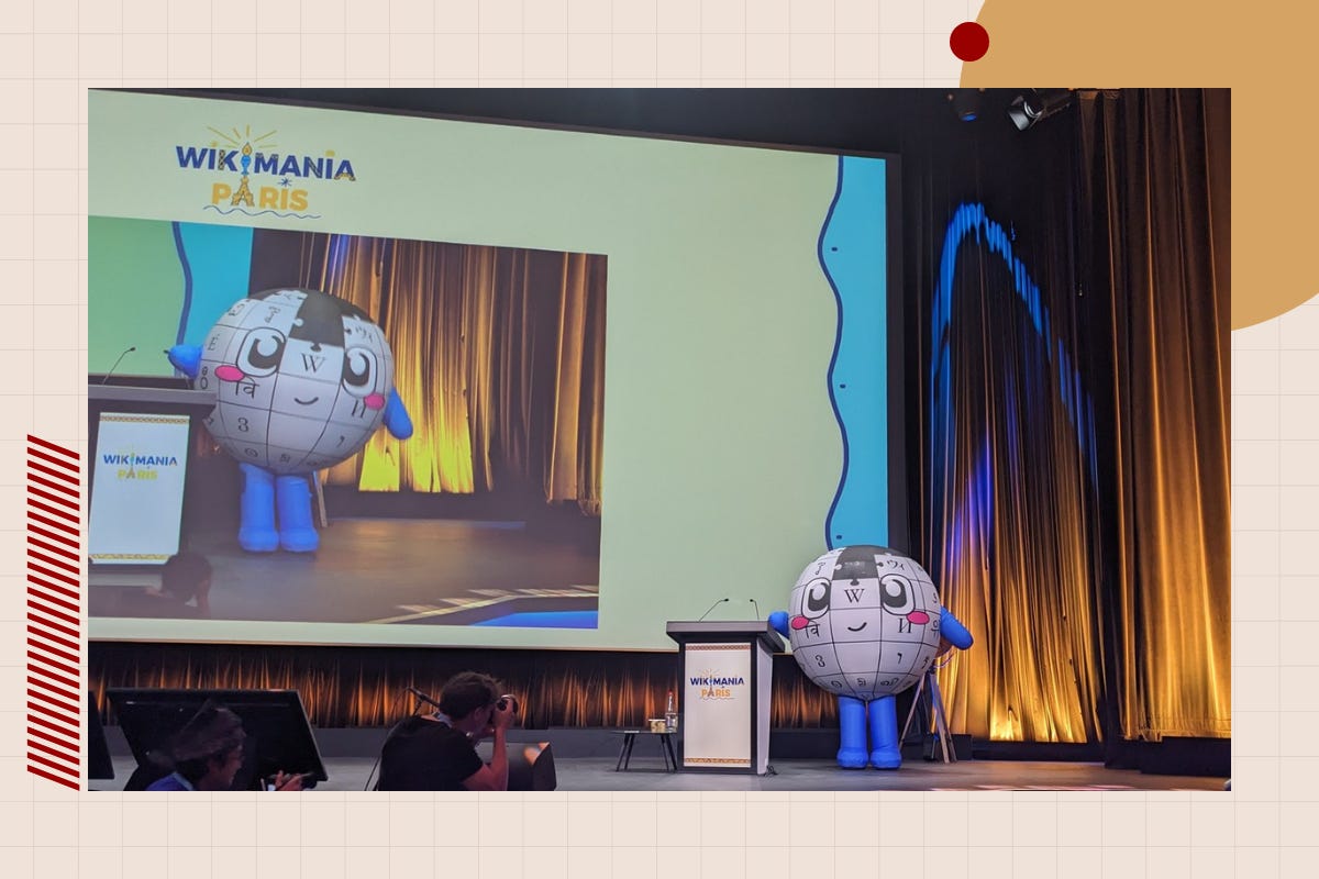 An inflatable Wikipedia puzzle-globe mascot with blue arms and legs stands onstage beside a Wikimania Paris podium, its own image displayed on the big screen behind it, as photographers shoot from below the stage. An inflatable Wikipedia puzzle-globe mascot with blue arms and legs stands onstage beside a Wikimania Paris podium, its own image displayed on the big screen behind it, as photographers shoot from below the stage.