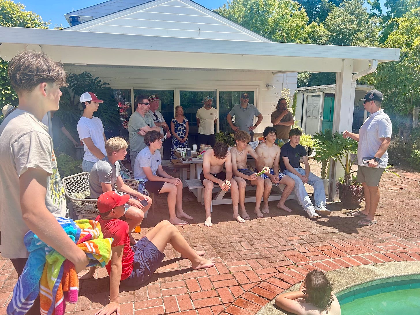 Coach Dan addressing the SRHS JV baseball team from behind the grill, mid-speech, surrounded by teens in swimsuits, donut floaties bobbing in the pool.