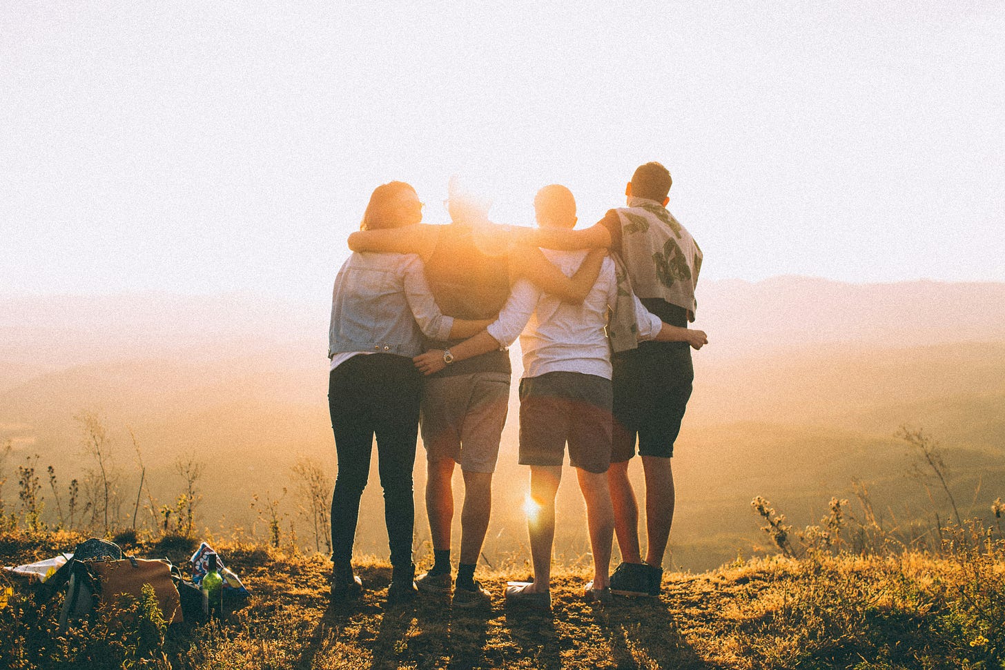 Photo taken from behind four people who are standing on a hill with their arms around each other, looking out at the view as the sun sets beyond a distant hill. Photo taken from behind four people who are standing on a hill with their arms around each other, looking out at the view as the sun sets beyond a distant hill.