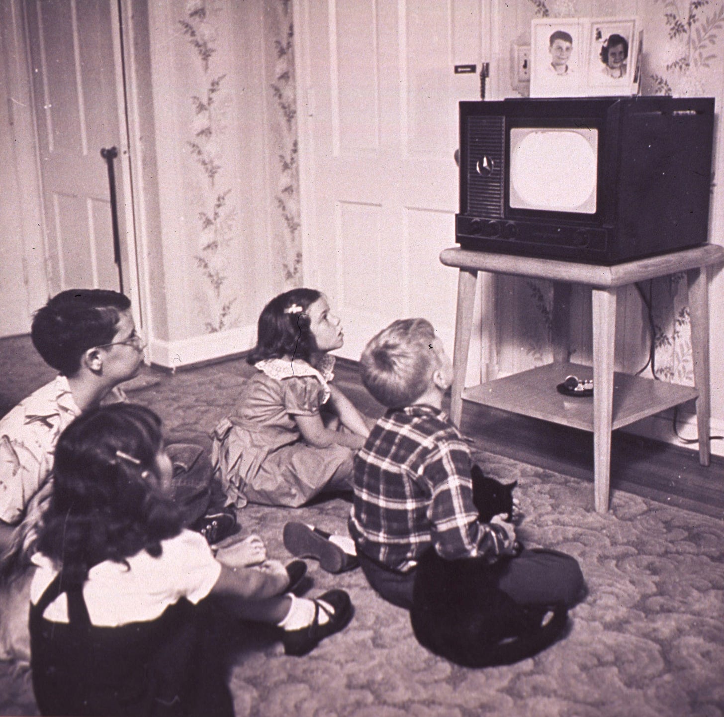 Four children from the mid-20th century sit on the floor watching a vintage television set, with family photos displayed on top, in a room with floral wallpaper.