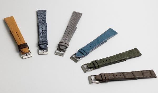 Six handmade watch straps made from unusual materials sit on a white table. From left: canvas in mustard, ostrich in blue, suede with ridges in gray, calfskin in blue, calfskin in green and alligator in brown.