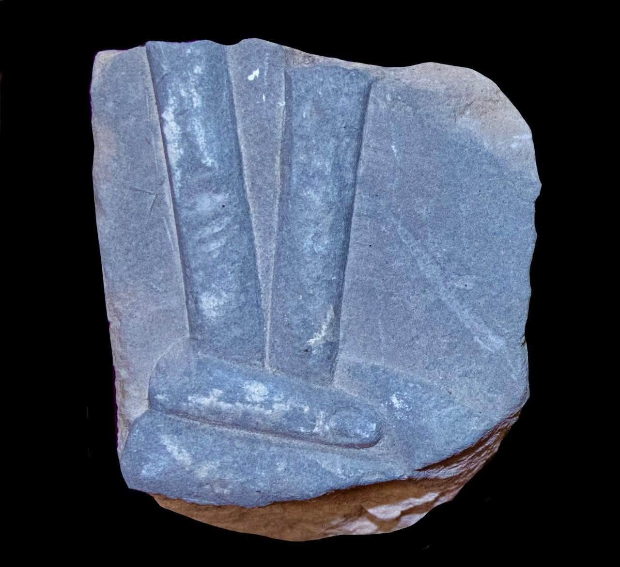 A carved fragment of dark stone from Tell el-Kharouba, showing part of a hand in raised relief.