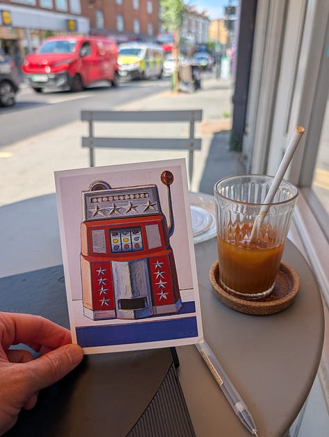 A table outside a bakery called Irene, with a black journal, pen, coffee in a glass cup and a postcard with pop art design.