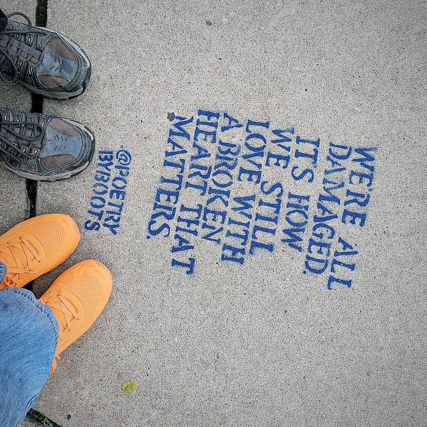We’re all damaged. It’s how we still love with a broken heart that matters. @PoetryByBoots stenciled in blue paint on pavement. Britne's orange shoes and jeans and Mike's brown shoes are visible.