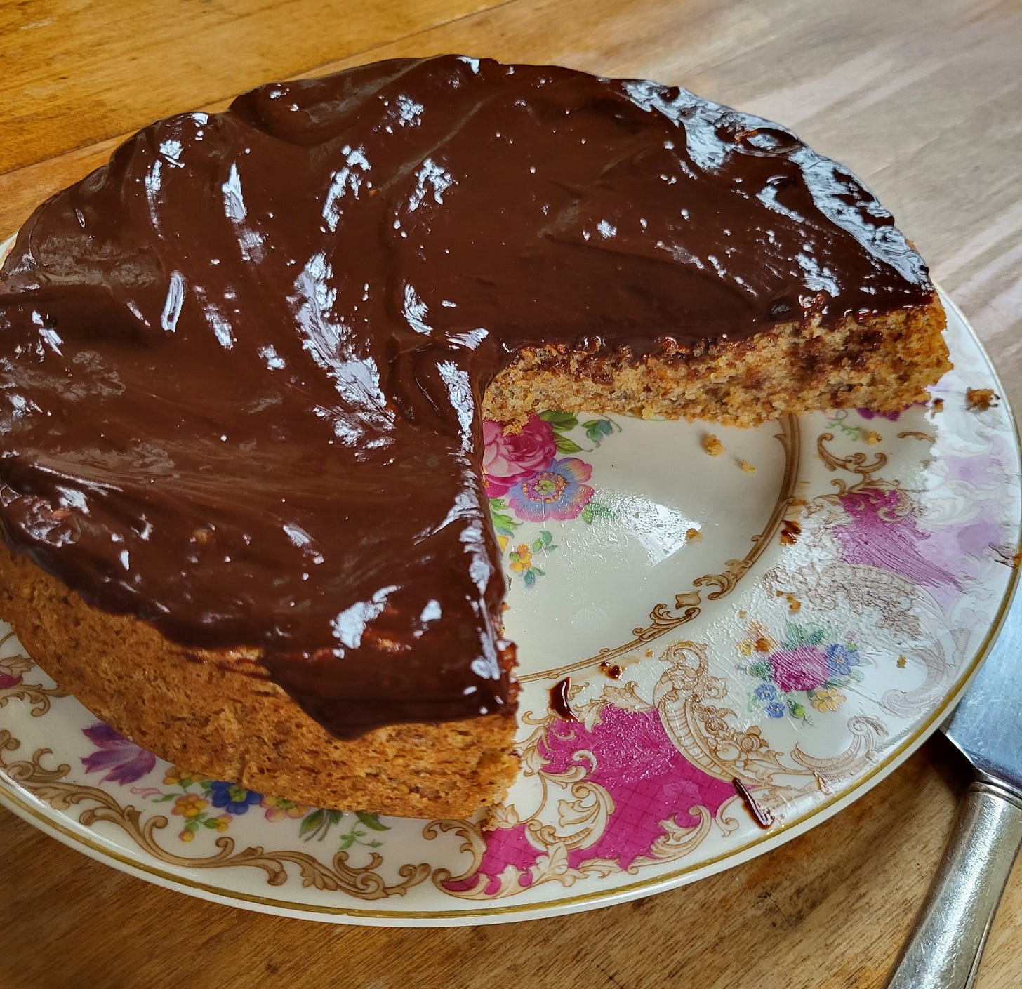 A hazelnut brown butter cake with very shiny chocolate ganache on a pink floral plate with a quarter missing