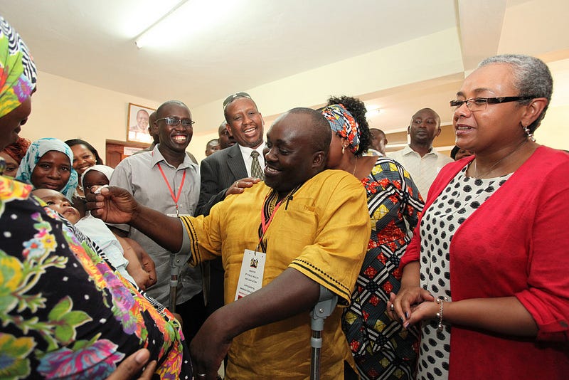 in the middle of a crowd, a smiling Black man using crutches administers an oral vaccine with a dropper to an infant in their mother's arms. everyone is smiling. in the middle of a crowd, a smiling Black man using crutches administers an oral vaccine with a dropper to an infant in their mother's arms. everyone is smiling.