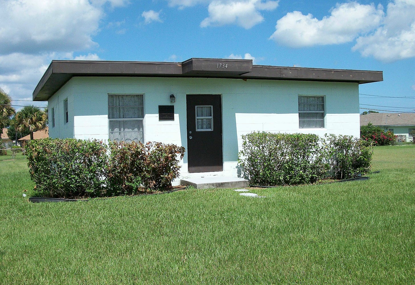 Photo of a small, boxy concrete cottage painted light green with a brown door and surrounding grass
