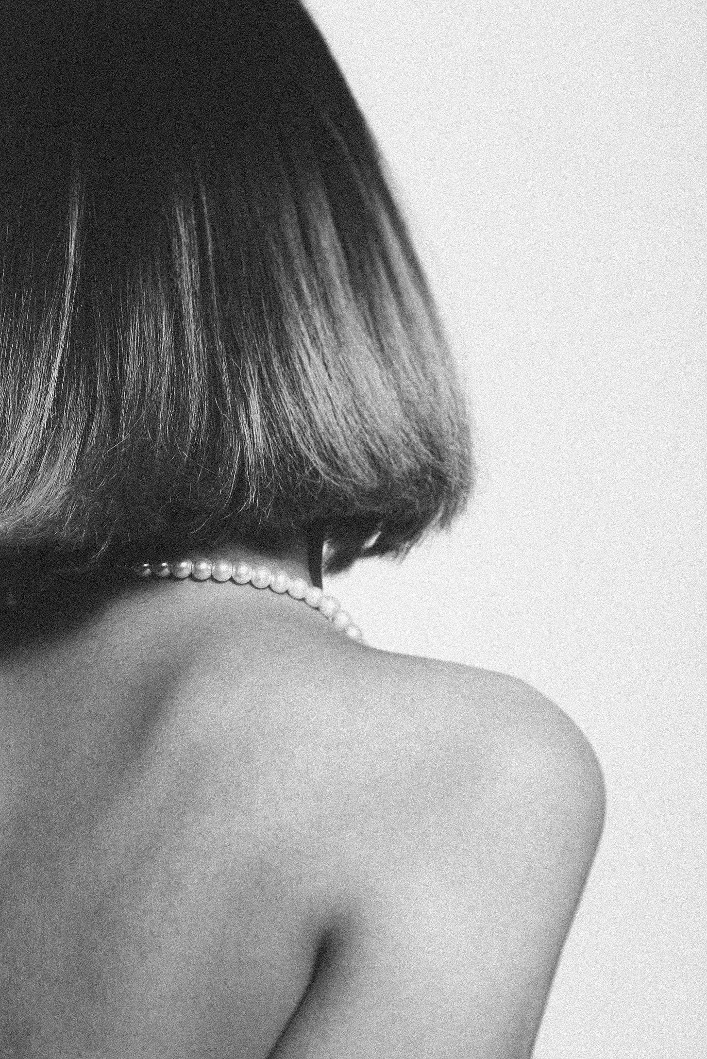 Back of a woman shows her bob, bare shoulders and a pearl necklace.