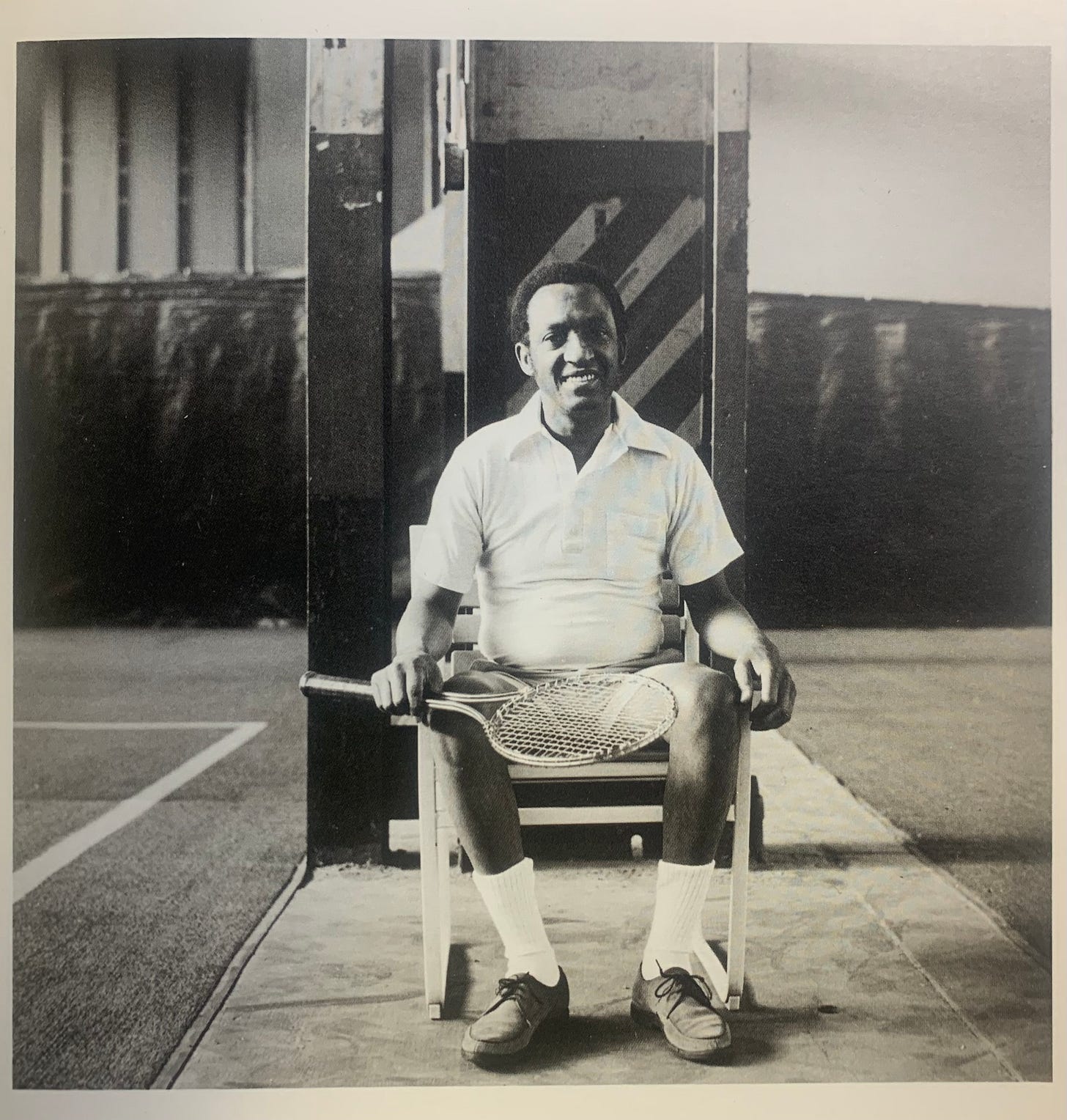 Tucker sitting in a chair looking forward at the camera smiling. He is holding a tennis racket in his left hand.