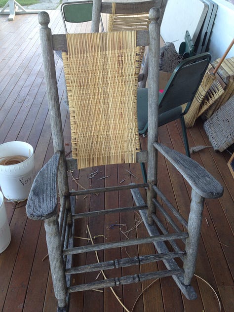 A rocker with a finished caned back, a group of caners show off their work, Jo in a blue t-shirt sitting in a finished rocker.