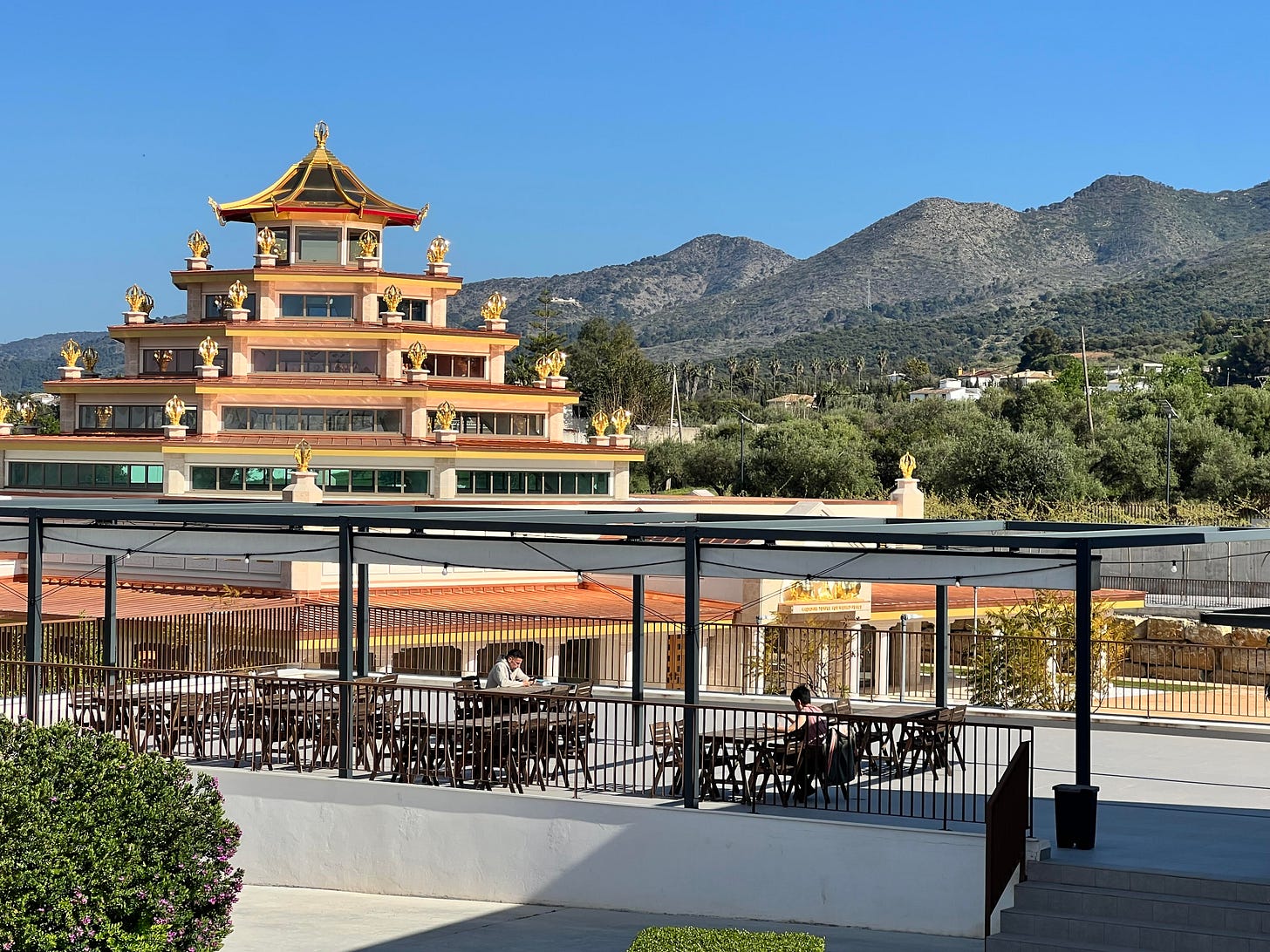 Open air dining terrace with wooden tables and chairs, the golden Buddhist temple pagoda rising behind it against a clear blue sky and mountain backdrop. Open air dining terrace with wooden tables and chairs, the golden Buddhist temple pagoda rising behind it against a clear blue sky and mountain backdrop.