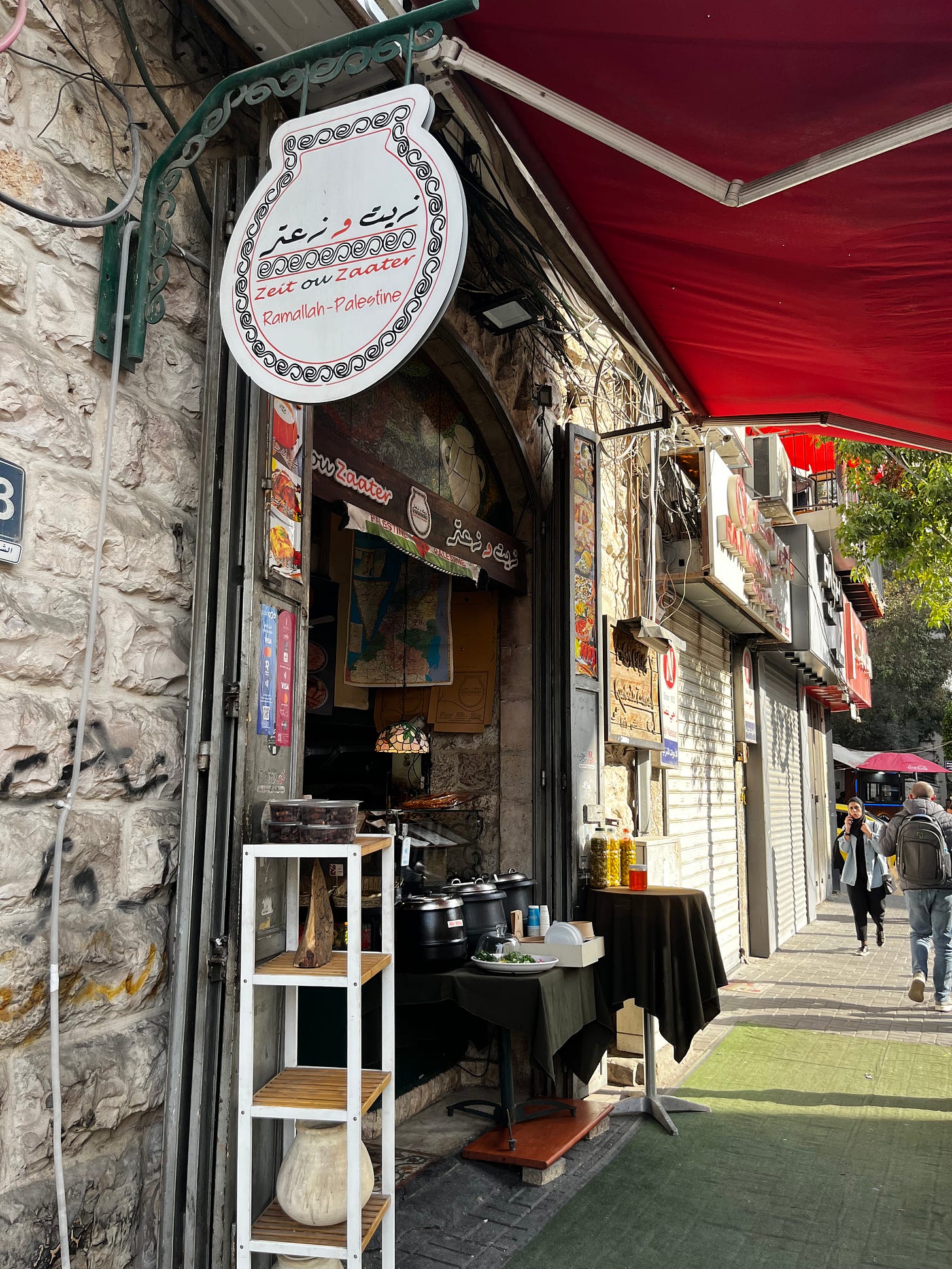 Street-level exterior of a stone-faced building with a red awning; a round café sign reads "Zeit & Zaater, Ramallah-Palestine" in Arabic and Roman script above an open arched doorway. Food warmers and display shelves line the entrance. Pedestrians walk the sidewalk beyond.