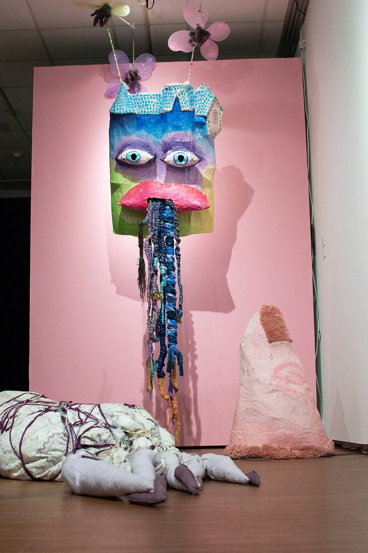 Installation shot of papiermache castle with eyes and mouth. From the mouth are multicolored yarn tendrils resembling roots, knots. A pink papiermache finger points at the castle. Fairies appear to hold the castle. The fairies have pastel wings and are in the shape of hands holding beaded string that connect to the back of the castle.