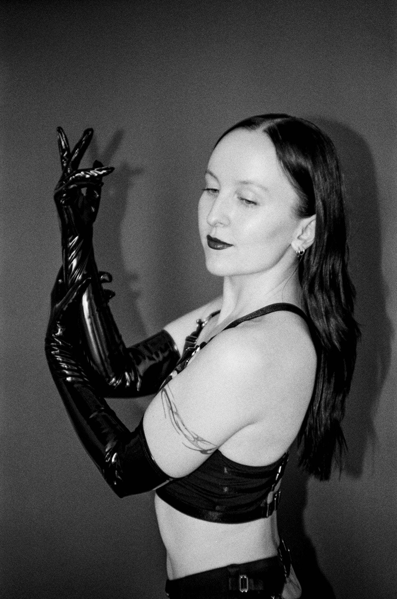 A grainy black and white photo of Evyn Silver, a white femme with long dark hair, posing with her hands raised in front of her. She wears long shiny black gloves, a strappy black outfit, and dark lipstick.