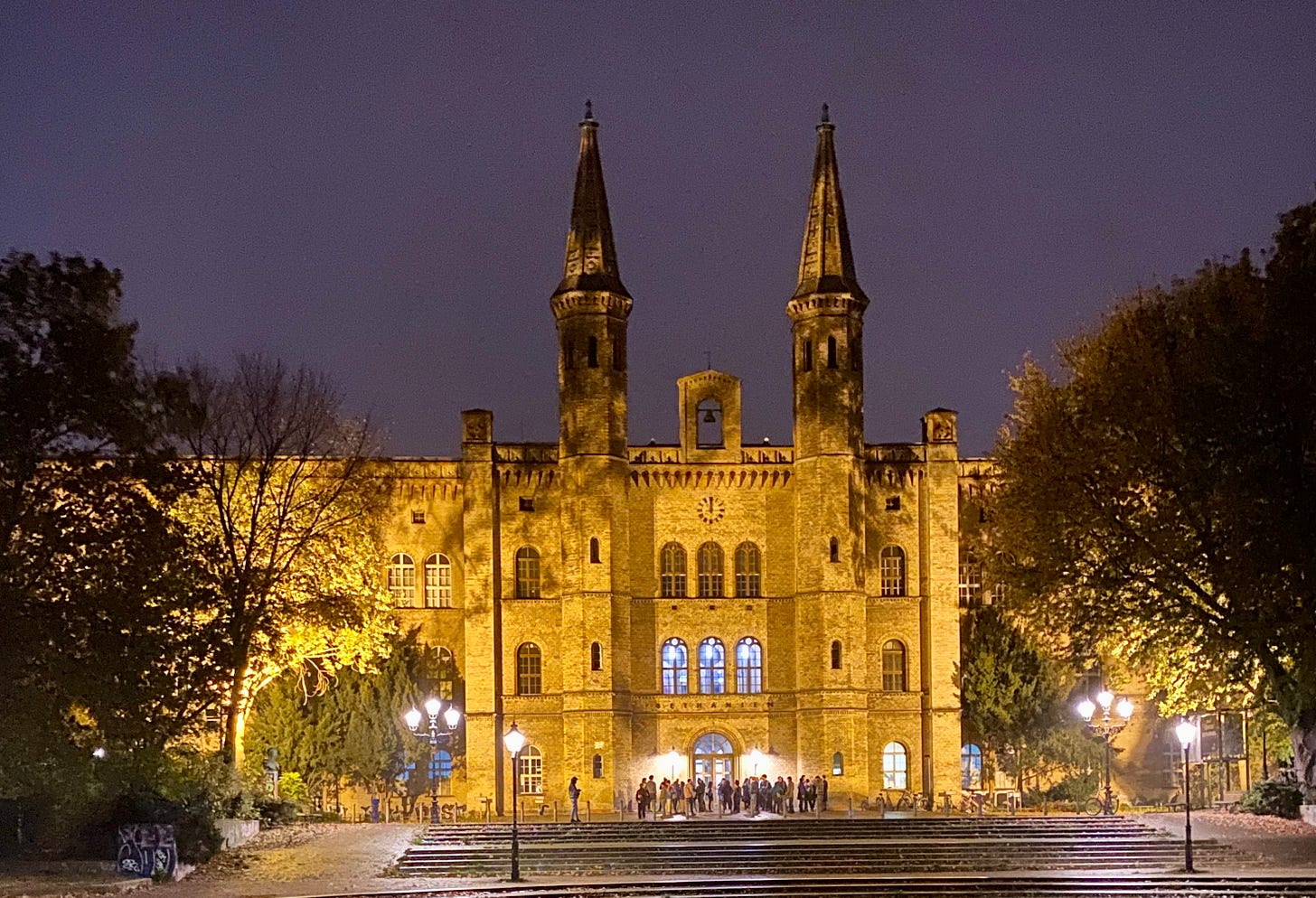 Edifício histórico iluminado à noite, com torres góticas, árvores ao redor e pessoas ao centro. Edifício histórico iluminado à noite, com torres góticas, árvores ao redor e pessoas ao centro.
