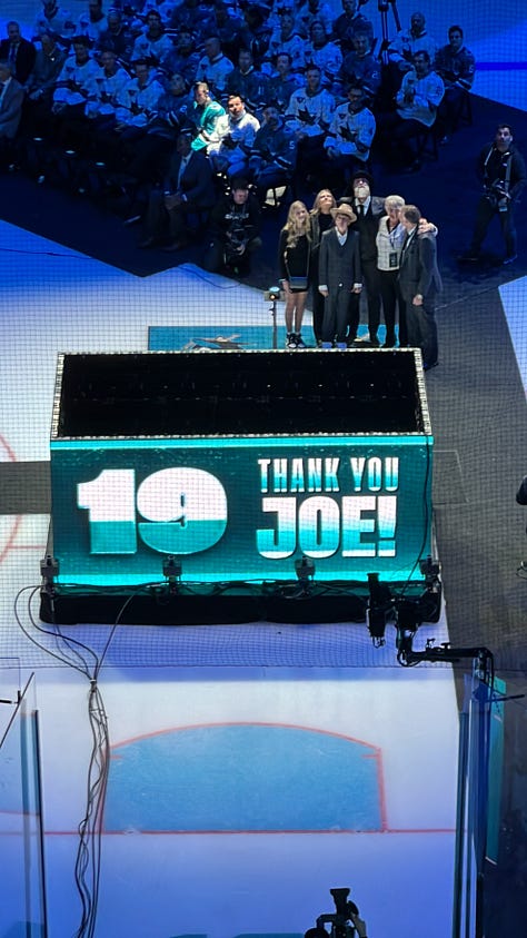 Photo 1: Image of woman in Sharks jersey smiling outside arena; Photo 2: Image of Joe Thornton ice sculpture; Photo 3: Image of Joe Thornton coming through Shark head on ice; Photo 4: image of two women smiling with ice rink behind them; Photo 5: Image of Shark head and chairs on ice for ceremony; Photo 6: Image of Joe Thornton on big screen; Photo 7: Image of Thank You Joe box where jersey was raised with family behind it; Photo 8: Image of ice rink with Shark head and players; Photo 9: Image of #12 and #19 jersey's hanging in the rafters of the arena