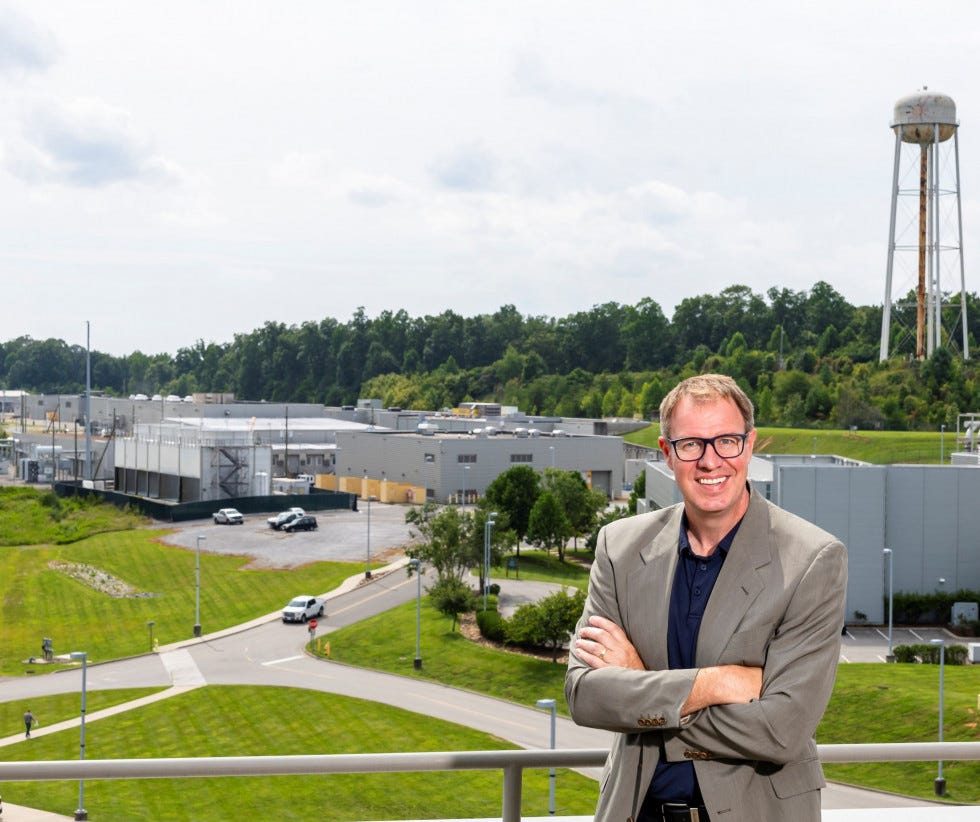 Person smiling with arms crossed in front of Oak Ridge National Lab, an industrial facility with a water tower in the background.