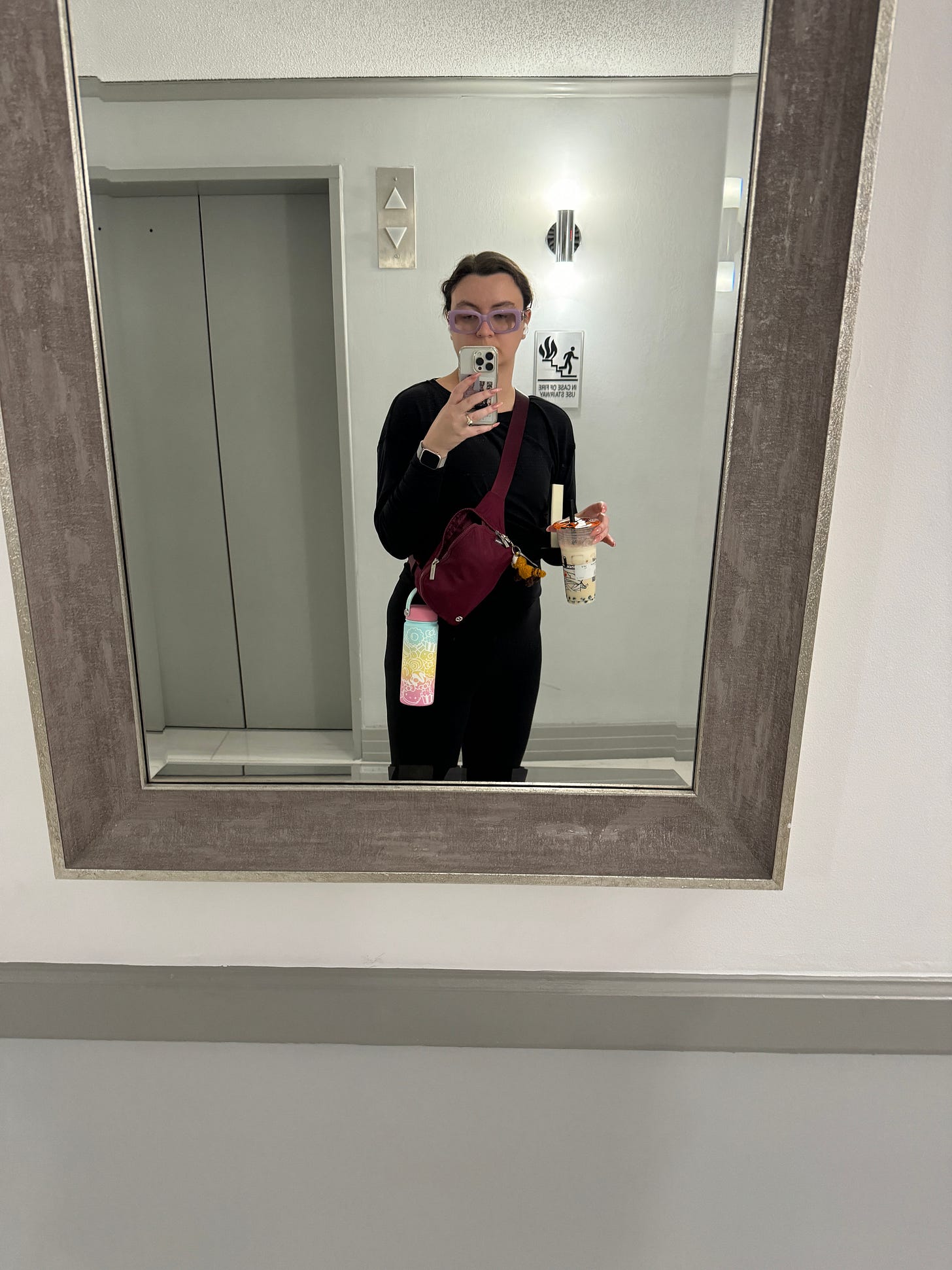 Woman in black pullover and bodysuit taking a mirror selfie at a bank of eleavators. Wearing a belt bag with a water bottle looped onto it, holding a boba tea, and with a book tucked under her arm. Woman in black pullover and bodysuit taking a mirror selfie at a bank of eleavators. Wearing a belt bag with a water bottle looped onto it, holding a boba tea, and with a book tucked under her arm.