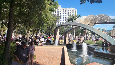 20251018_114518.jpg: A close-up of a hand-drawn protest sign reading "NO KINGS!!" and "NO WEENIES ALLOWED," featuring a crossed-out Burger King logo and a drawing of SpongeBob SquarePants.  20251018_113815.jpg: A wide shot of a large, sunny protest in a public plaza, showing crowds gathered around a modern arched sculpture and a water fountain.  20251018_113805.jpg: A crowd of protestors gathered on a brick plaza under a canopy of trees, with some people holding American flags and various signs.  20251018_110151 - Copy.jpg: A person in an inflatable blue pterodactyl costume stands at the protest, next to a person in a wheelchair holding a sign that reads, "DECLARE INDEPENDENT. FLUSH THE RED/BLUE WAR!"  20251018_113823.jpg: A wide view of the protest centered on a large, modern arched sculpture over a fountain, with a large crowd and many signs visible in the background.