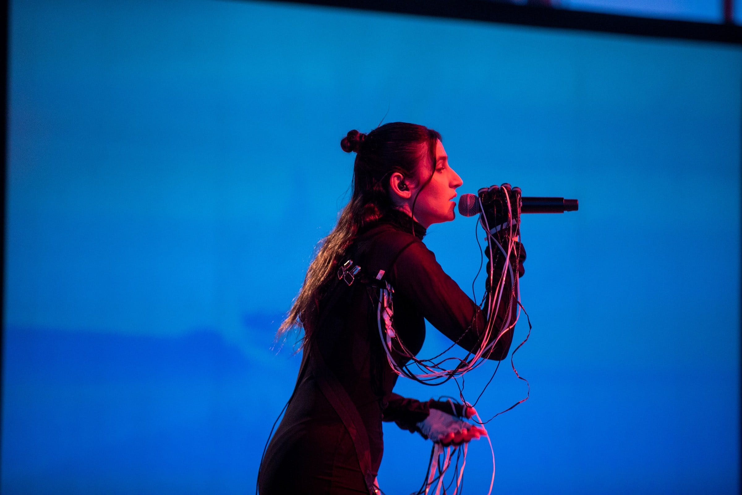 A singer in profile at a wired microphone against a glowing blue projection wall, lit magenta from the side A singer in profile at a wired microphone against a glowing blue projection wall, lit magenta from the side