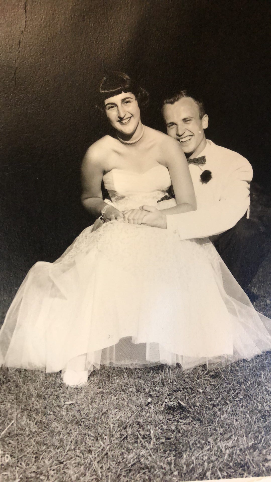 A young couple posing in formal wear; she is in a flowing white dress seated, perhaps, on his knee who is squatting behind with on hand in her hand on her lap, wearing a white jacket. Both smile broadly.