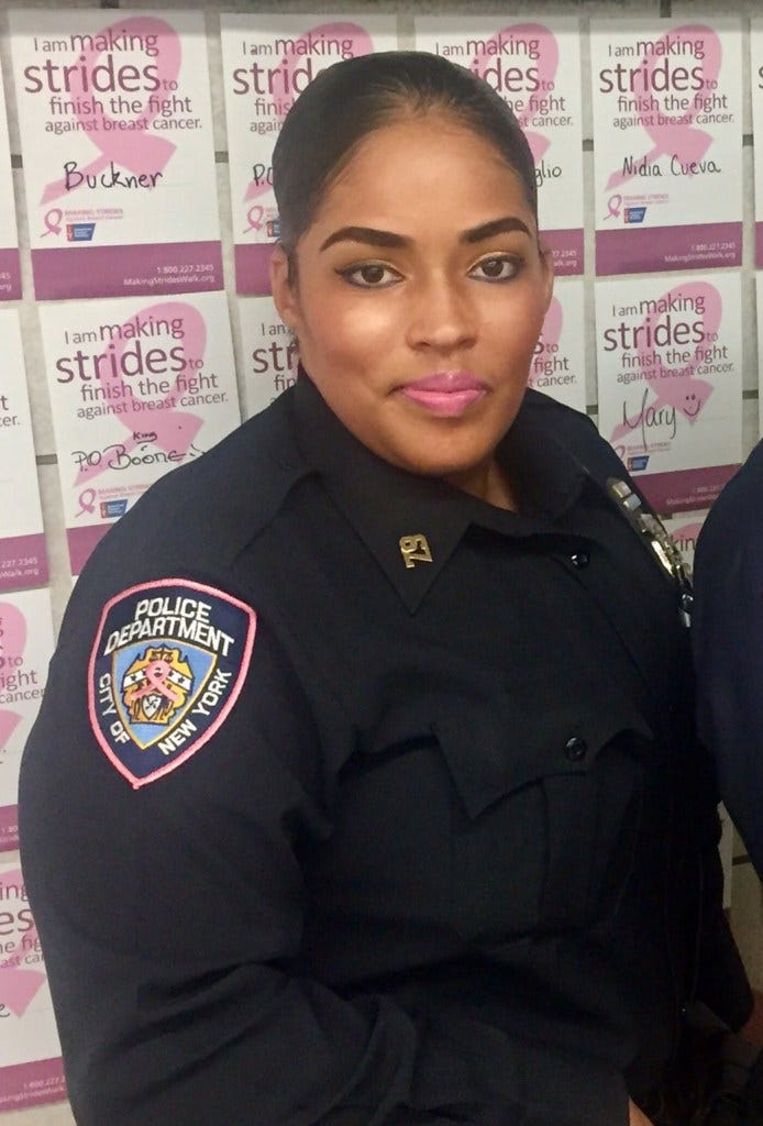 NYPD Detective Alicia Stone in uniform with breast cancer awareness posters in the background.