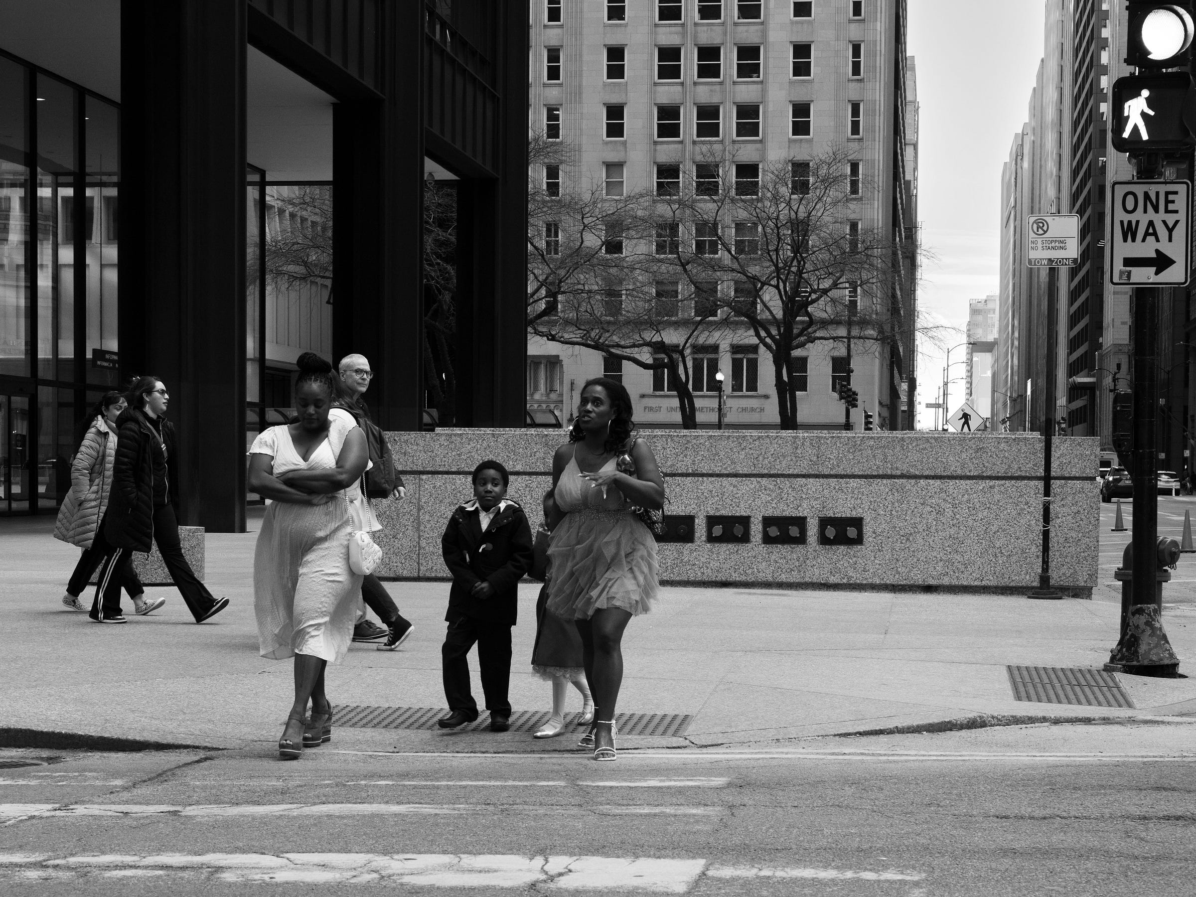 Black and white photograph of three people crossing a city street, dressed in formal attire. A woman in a flowing dress walks confidently, gesturing as she speaks, while a young boy in a suit follows closely beside her. Another woman in a white dress walks with arms crossed. In the background, pedestrians in casual clothing pass by modern office buildings, contrasting the subjects’ elegant outfits with the everyday cityscape. Black and white photograph of three people crossing a city street, dressed in formal attire. A woman in a flowing dress walks confidently, gesturing as she speaks, while a young boy in a suit follows closely beside her. Another woman in a white dress walks with arms crossed. In the background, pedestrians in casual clothing pass by modern office buildings, contrasting the subjects’ elegant outfits with the everyday cityscape.