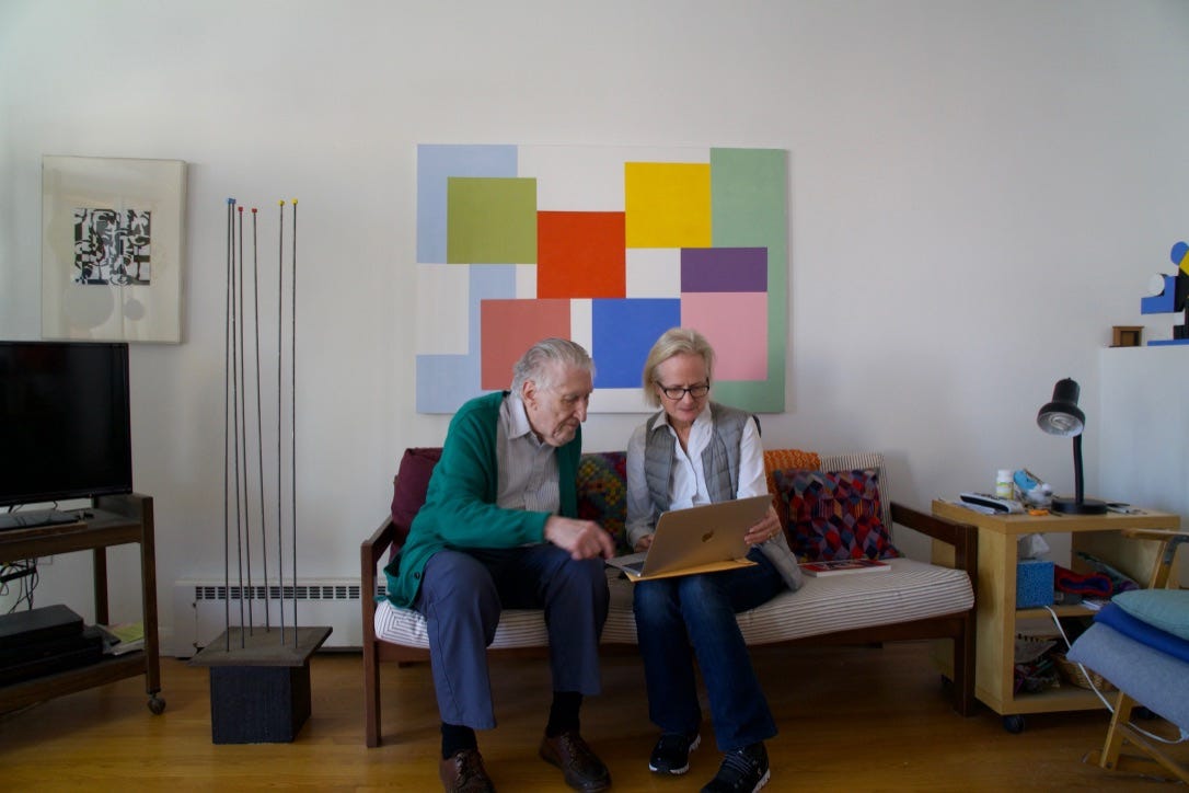 man and woman sitting on a couch looking at a laptop together, pointing. A brightly colored geometric painting hangs on the wall overhead and other art works surround them.