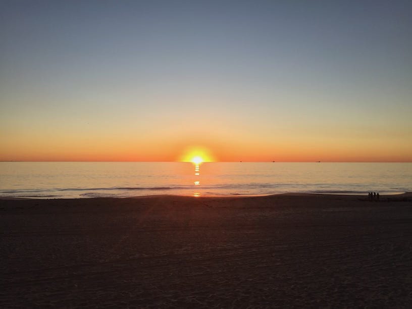 A serene sunset over the ocean with the sun low on the horizon, casting a golden reflection across the calm water. The sky fades from warm orange near the horizon to deep blue above, with a sandy beach in the foreground and a few people visible on the right.
