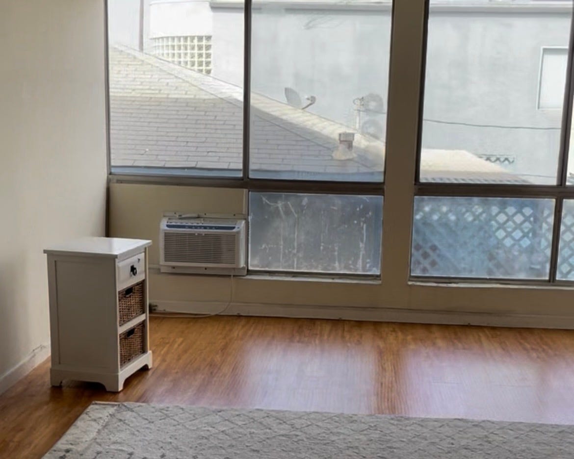 An empty room in a small Los Angeles apartment with wood floors, a window air conditioning unit, and large windows looking onto the roof of the neighboring house and overhead power lines. An empty room in a small Los Angeles apartment with wood floors, a window air conditioning unit, and large windows looking onto the roof of the neighboring house and overhead power lines.