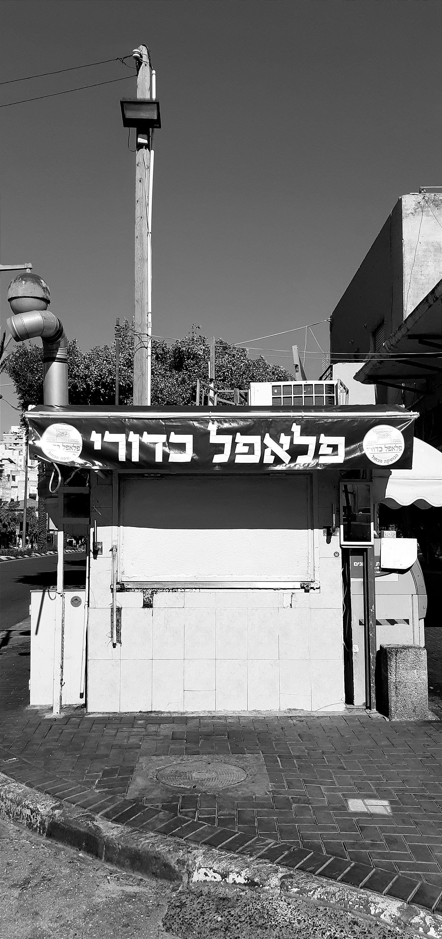 A small falafel stand photographed straight-on, its metal service shutter closed. A large blue banner above reads "Falafel Kaduri" in white Hebrew lettering. The stand is a freestanding tiled structure at a street corner; residential buildings and trees are visible in the background. The image is in black and white.