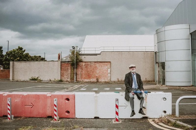 Nev Clay sits on concrete barriers in front of an abandoned car park. He is holding a closed umbrella.