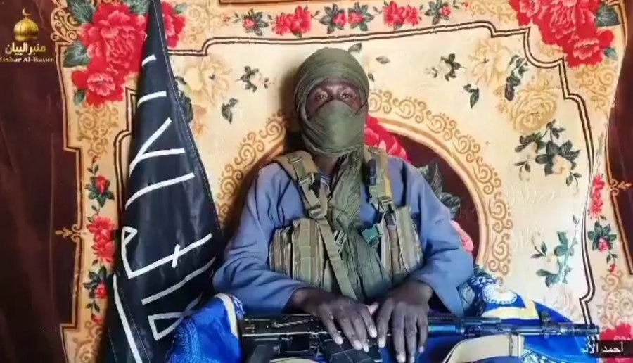 A man wearing a green turban and face mask sits in front of a large black flag with white Arabic script and a gold emblem. He is dressed in tactical vest and clothing holding a rifle across his lap. The background features ornate floral designs in red and gold with circular patterns.