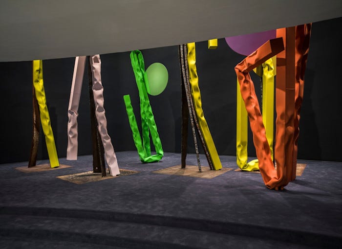 Tall, crumpled metal sculptures in bright yellow, pink, green and orange stand on a dark carpeted floor with circular coloured panels behind them.