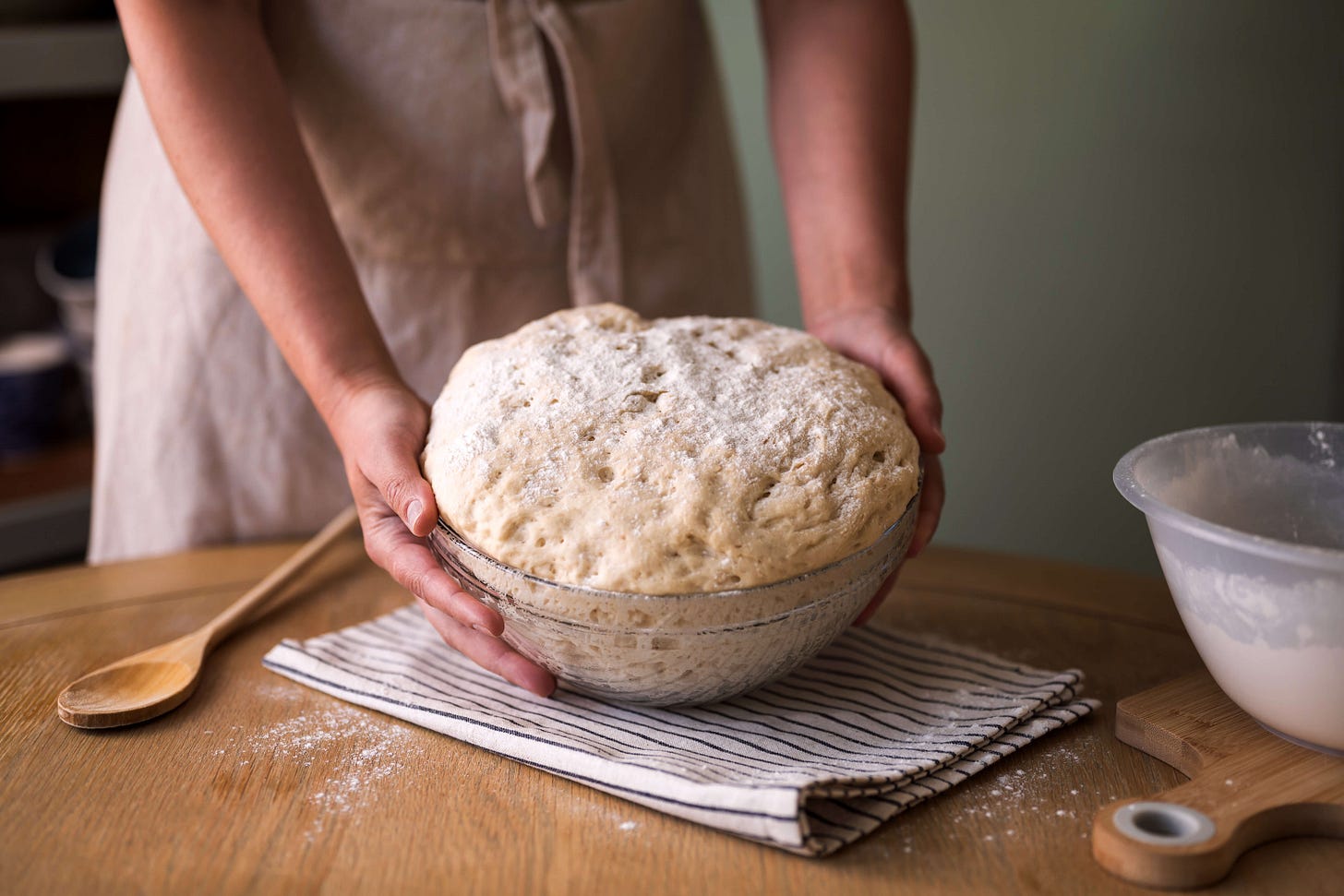 Die Hände einer Frau in Schürze halten eine Schüssel mit aufgegangenem Sauerteig auf einem Holztisch. Daneben liegen ein Holzlöffel, ein gestreiftes Tuch und eine weitere Teigschüssel – Vorbereitung für das Brotbacken.