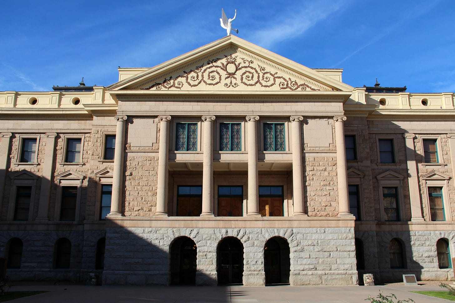 The front facade of the historic Arizona State Capitol building, a tan stone structure with four tall columns, a decorative triangular pediment, and a winged "Winged Victory" figure mounted at its peak against a clear blue sky.
