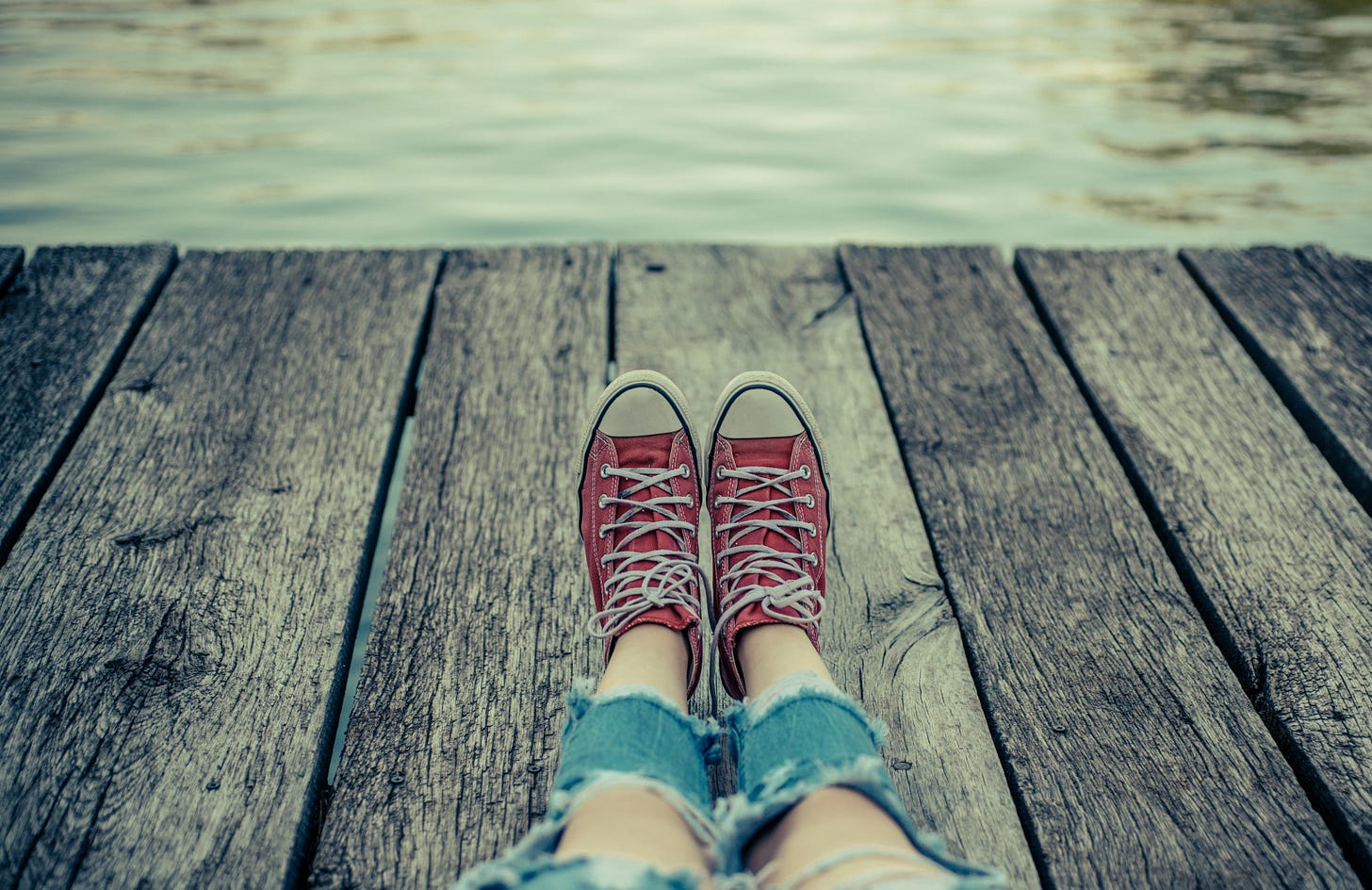POV of person sitting on a boardwalk by the water. Their legs are extended in front of them; they’re wearing ragged jeans with the knees cut-out, and red sneakers.