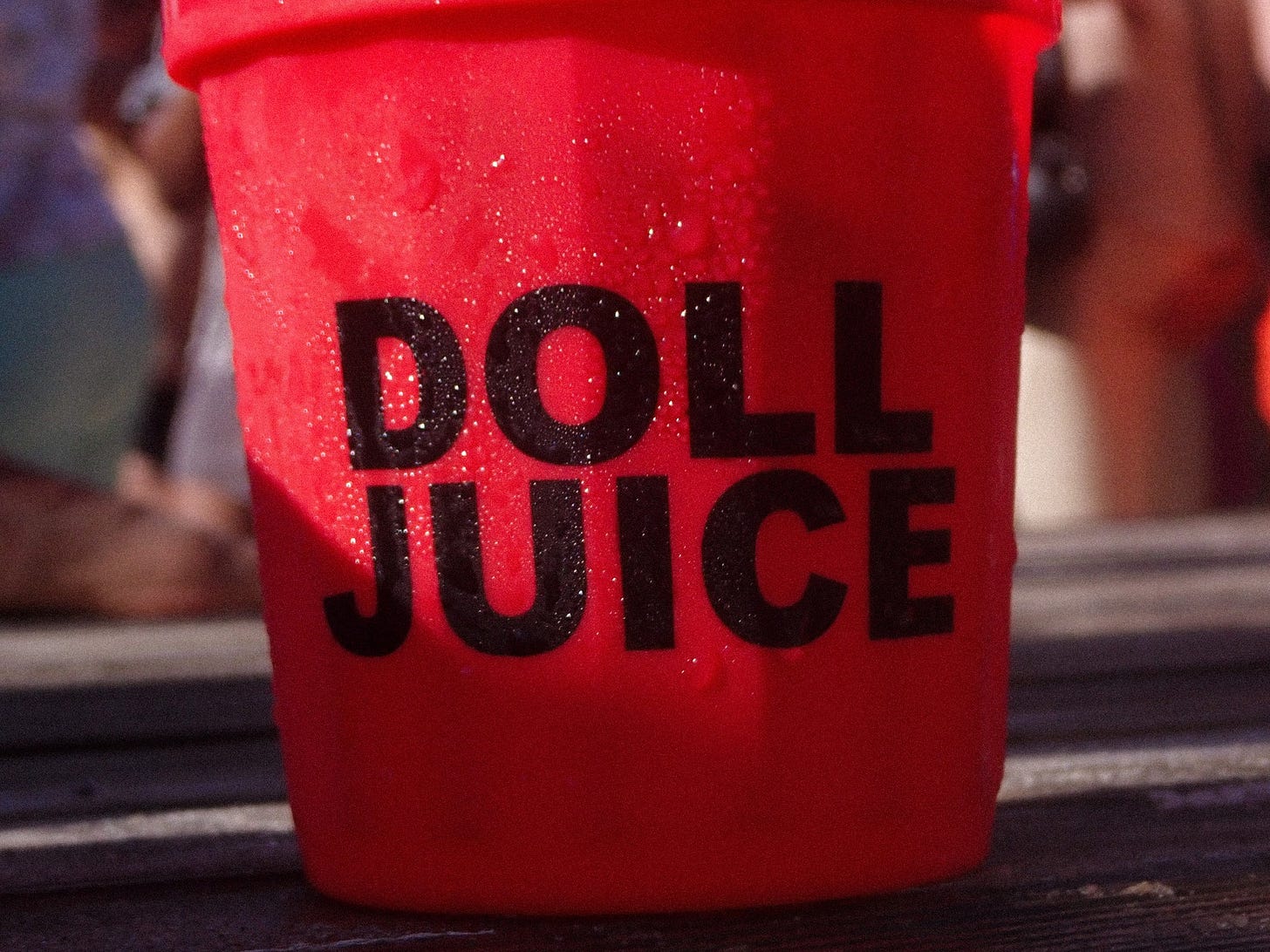 A red plastic cup with "DOLL JUICE" written in bold black letters, glistening with condensation under bright sunlight.