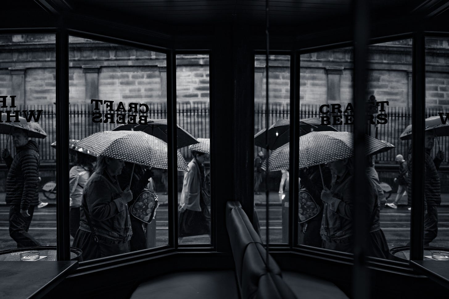 Figures under umbrellas seen through the mirrored glass panels of a shelter, their reflections doubled against the street beyond. Figures under umbrellas seen through the mirrored glass panels of a shelter, their reflections doubled against the street beyond.