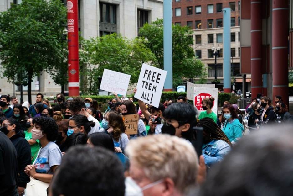 People stand outside in masks with a sign in the center that says Free Them All!