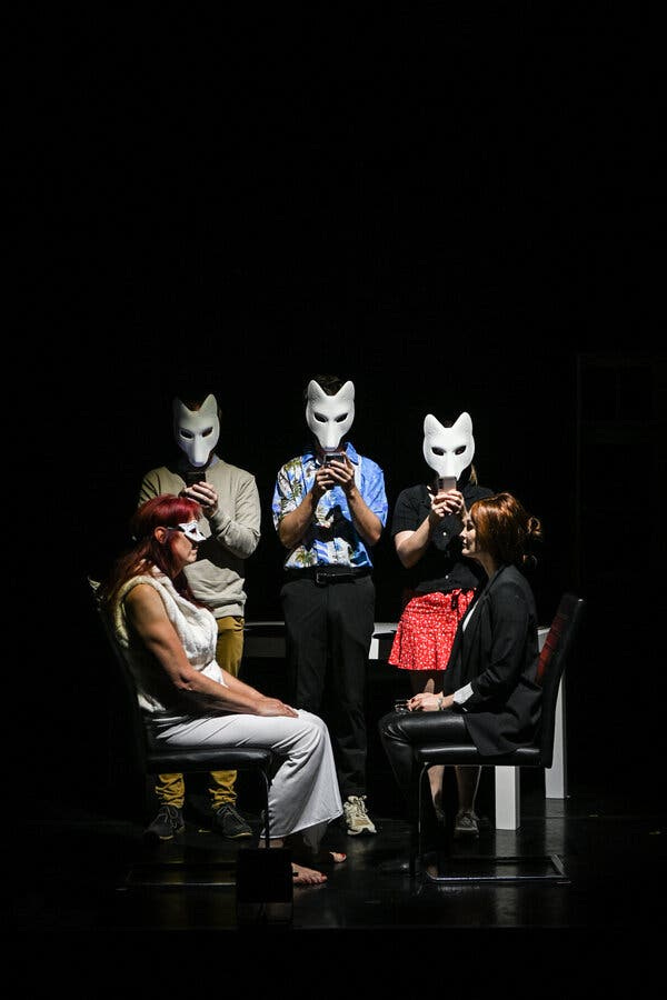 Two women sit facing each other on a darkened stage; behind them, three actors wearing white masks stand in a row.